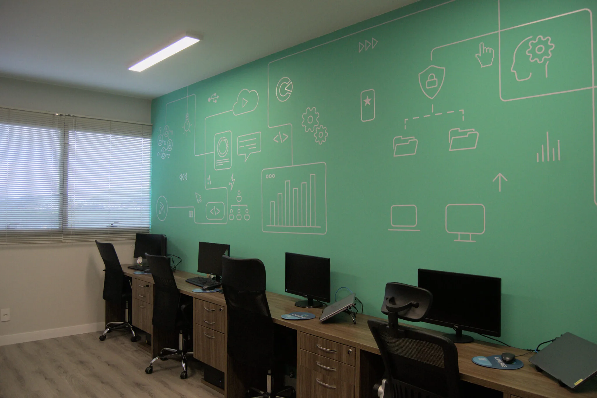 Sala de escritório com estações de trabalho organizadas lado a lado, cada uma equipada com monitor, teclado e cadeira ergonômica