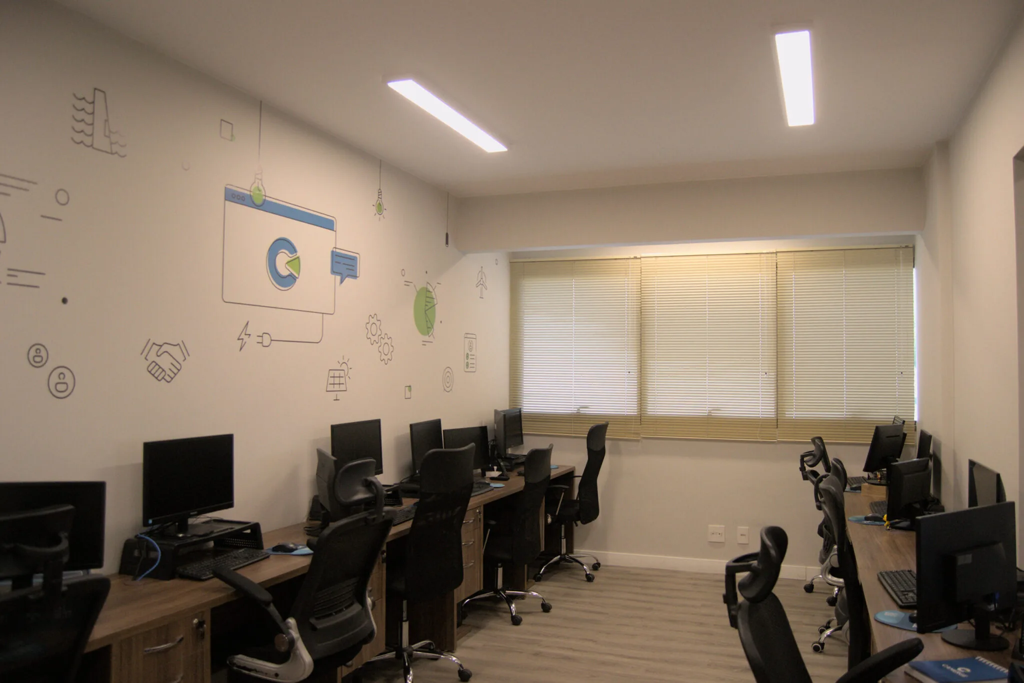 Sala de escritório com estações de trabalho organizadas nas duas paredes laterais, cada uma com monitor, teclado, CPU e cadeiras ergonômicas