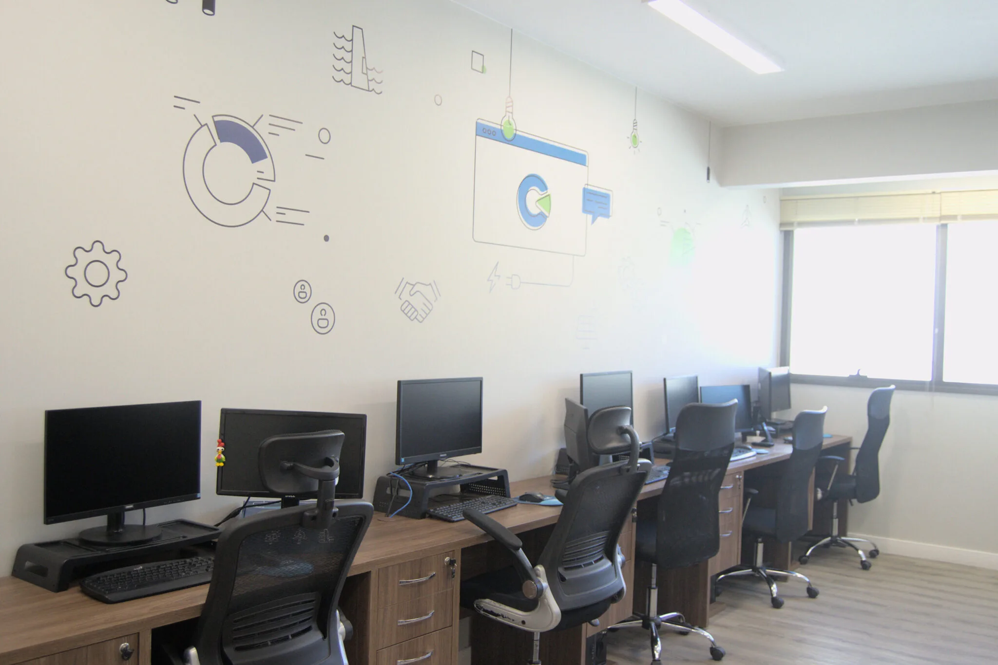 Sala de escritório com várias estações de trabalho alinhadas em uma única parede, cada uma com monitor, teclado e cadeira ergonômica