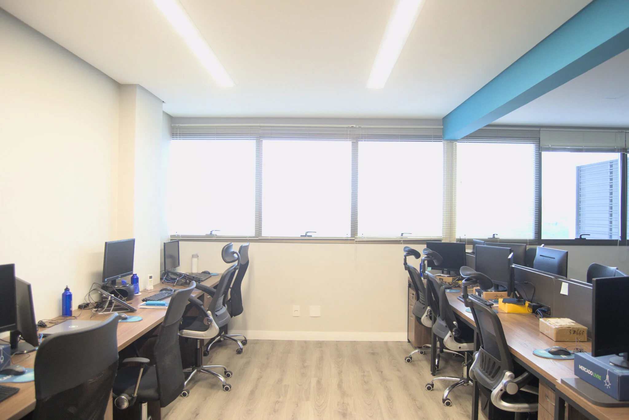 Sala de escritório com duas fileiras de mesas paralelas, cada uma equipada com monitores, teclados e cadeiras ergonômicas.