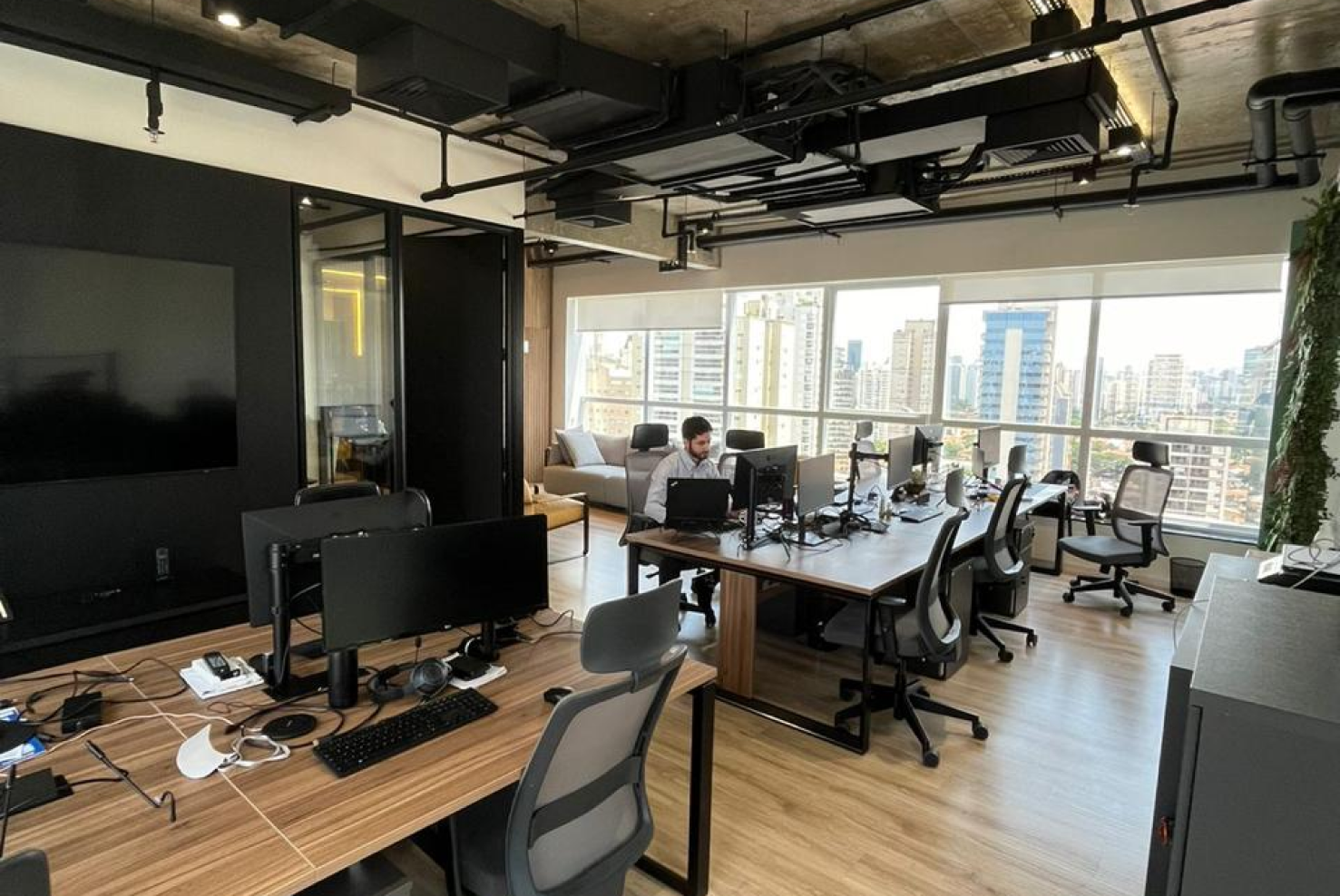 Escritório moderno com estações de trabalho, cadeiras ergonômicas, computadores e uma grande janela com vista para prédios da cidade ao fundo. Um homem trabalha em uma das mesas."
