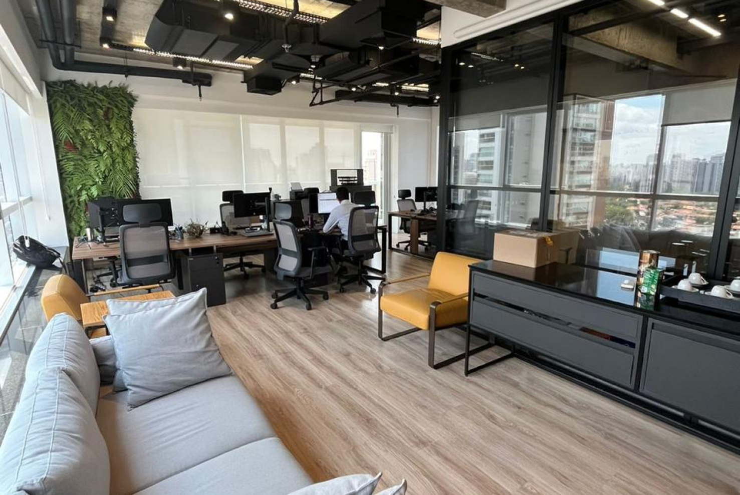 Escritório moderno com piso de madeira clara, estações de trabalho com cadeiras ergonômicas e computadores, uma parede com jardim vertical ao fundo